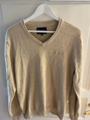 Beige stickad tröja från Bondelid - Snygg beige stickad tröja från Bondelid med V-ringning och diskret broderad logga på bröstet. Tröjan har långa ärmar och ribbade muddar vid ärmslut och nederkant. Perfekt för lager-på-lager och enkel att matcha med jeans eller chinos.