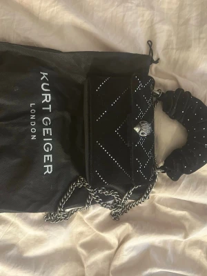 Svart handväska från Kurt Geiger - Snygg svart handväska från Kurt Geiger med silverfärgade kedjedetaljer och dekorativa små stenar i rutmönster. Väskan har ett vadderat handtag med scrunchie-look och ikonisk örndetalj på locket. Perfekt för att lyfta din outfit.