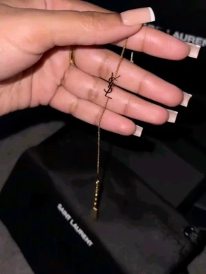 Guldigt YSL armband - Snyggt och stilrent armband från Saint Laurent i guldtonad metall. Armbandet har en tunn kedja och ett ikoniskt YSL-logotyp-hänge i mitten. Perfekt accessoar för dig som gillar lyxiga detaljer och minimalistisk design.