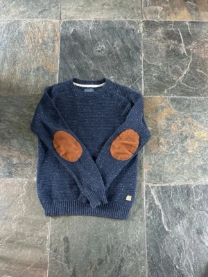 Mörkblå stickad tröja från Blend - Mörkblå stickad tröja från Blend med vita småprickar och bruna mockadetaljer på armbågarna. Tröjan har rund halsringning, ribbade muddar och en klassisk passform. Perfekt för dig som gillar en stilren och avslappnad look.