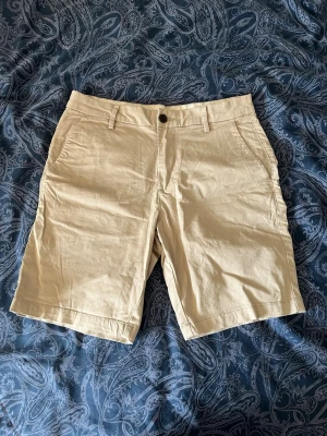 Beige chinoshorts med fickor - Snygga beige chinoshorts med klassisk passform och raka ben. Shortsen har två sidofickor, bälteshällor och stängs med knapp och dragkedja. Tillverkade i ett mjukt bomullsmaterial som ger en avslappnad men stilren look.