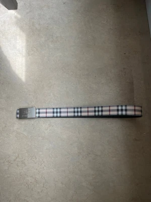 Burberry bälte - Snyggt bälte från Burberry med klassiskt rutmönster i vitt, svart, rött och blått. Bältet har ett stilrent spänne i silverfärgad metall med Burberry-logga. Perfekt accessoar för att lyfta din outfit.