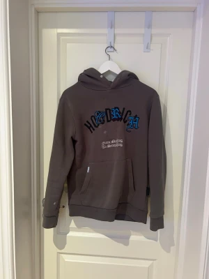 Grå hoodie från HOODRICH - Snygg grå hoodie från HOODRICH med blå och svart text på bröstet och stor tryckt text på ryggen. Klassisk känguruficka framtill och huva med dragsko. Perfekt för en streetwear-look och riktigt skön att bära.(fläckarna är borta)
