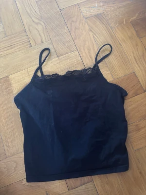 Svart kort linnetopp med spetskant - Svart linnetopp med tunna axelband och fin spetsdetalj längs urringningen. Enkel och stilren modell som passar perfekt till jeans eller kjol. Mjuk och bekväm känsla.