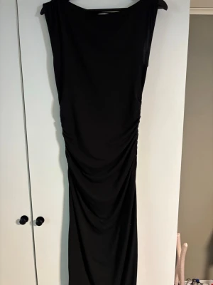 Svart långklänning från Kappahl XS - En stilren svart långklänning från Kappahl i storlek XS. Klänningen har en enkel, elegant design med rynkade detaljer längs sidorna och är ärmlös. Perfekt för dig som gillar en klassisk look med modern twist.