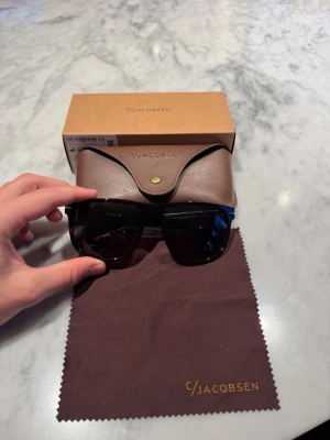 C/Jacobsen bruna solglasögon - Snygga solglasögon från C/Jacobsen med mörkbruna, fyrkantiga bågar i plast och mörka linser. Kommer med ett brunt fodral i läderlook och putsduk med logga. Perfekt accessoar för att lyfta din stil.