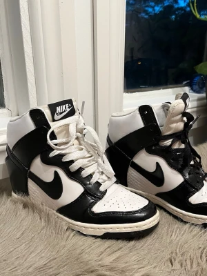 Nike svartvita high top wedgeboots - Säljer ett par snygga svartvita high top boots med inbyggd klack från Nike med klassisk swoosh-logga på sidorna. Skorna har vit bas med svarta detaljer och snörning hela vägen upp. Perfekta för dig som gillar streetstyle och sportig look.