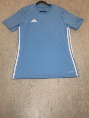 Blå sporttröja från Adidas - Snygg ljusblå sporttröja från Adidas i slim fit-modell. Tröjan har korta ärmar, vita ränder längs sidorna och är tillverkad i ett lätt och snabbtorkande material. Perfekt för träning eller match.