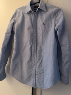Ljusblå Ralph Lauren skjorta - Klassisk ljusblå skjorta från Polo Ralph Lauren med broderad logga på bröstet. Skjortan har lång ärm, knappar framtill och en tidlös passform. Perfekt för en clean och stilren look.