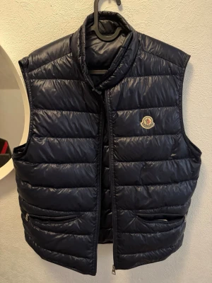 Mörkblå dunväst från Moncler - Snygg mörkblå dunväst från Moncler med hög krage och dragkedja framtill. Västen har två fickor med dragkedja och det klassiska Moncler-logotypen på bröstet. Perfekt för lager-på-lager och riktigt skön att bära under kyliga och varma dagar, den är väldigt tun och e köpt från merchsweden, kom skriv om ni är intresserade.