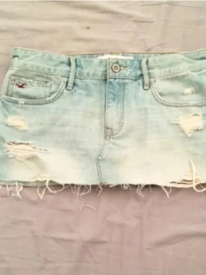 Ljusblå jeanskjol från Hollister - Säljer en ljusblå jeanskjol från Hollister med slitna detaljer och fransig nederkant. Kjolen har klassiska fickor fram och bak samt knapp och dragkedja framtill. Perfekt för dig som gillar en avslappnad och trendig stil. Säljer då den blivit lite tajt på mig