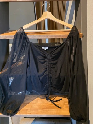 Svart kort top med ballongarmar från bubbleroom  - Trendig svart blus med transparenta, långärmade puffärmar och snörning framtill. Blusen har en croppad passform och urringning som ger en cool och modern look. Perfekt att styla med höga jeans eller kjol. Storlek L 
