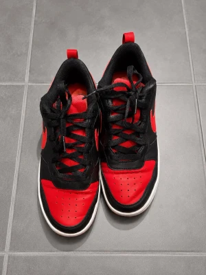 Nike svart-röda sneakers låga - Snygga sneakers från Nike i svart och rött med klassisk Swoosh på sidan. Skorna har lågt skaft, svart ovandel i läder och röda detaljer på tå, sula och logga. Perfekta för dig som vill ha en sportig och trendig look.