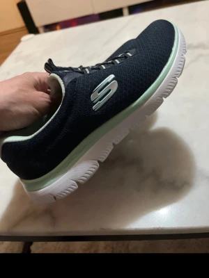 Svarta Skechers sneakers med vit sula - Snygga svarta Skechers sneakers med vit sula och mintgröna detaljer. Ovandelen är i meshmaterial för extra andningsförmåga och komfort. Skorna har elastiska snören och Skechers-logga på sidan. Perfekta för en sportig och avslappnad stil. Storlek 36