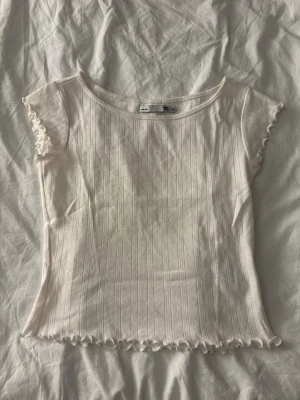 Vit ribbad topp från 157 - Supersöt vit ribbad topp från 157 med korta ärmar och volangkant vid ärmslut och nederkant. Tunt och mjukt material, perfekt till sommaren. Enkel och stilren design med diskreta lodräta ränder.