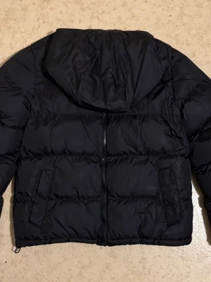 Svart pufferjacka från Hoodrich - Svart pufferjacka från Hoodrich med huva och dragkedja framtill. Jackan har två snedställda fickor och quiltad design som ger en schysst streetkänsla. Perfekt för kalla dagar och enkel att matcha med olika outfits.