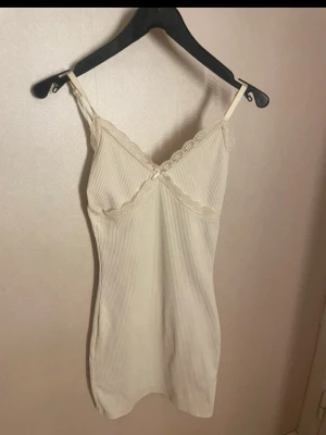 Beige ribbad klänning med spetsdetaljer - Söt beige ribbad klänning med smala axelband och spetsdetaljer längs urringningen. Klänningen har en liten rosett framtill och är figurnära med mjukt material. Perfekt för en romantisk look.