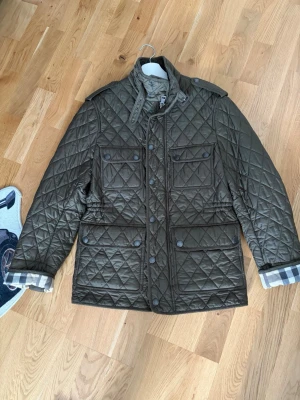 Olivgrön quiltad jacka från Burberry - Snygg olivgrön quiltad jacka från Burberry med klassiskt rutigt foder i ärmsluten. Jackan har fyra fickor framtill med lock och knappar, axelklaffar och hög krage med spänne. Perfekt för dig som vill ha en stilren och tidlös look.