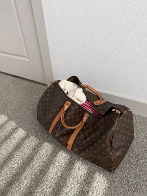 monogram resväska - Säljer en klassisk resväska i brunt monogrammönster med detaljer i cognacsfärgat läder och guldfärgade metalldelar. Väskan har dubbla handtag och loggor över hela ytan. Perfekt för weekendresor och snygg att bära med sig.