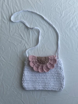 Virkad vit handväska med rosa lock - Söt handväska i virkat vitt garn med axelrem och ett dekorativt lock i rosa och beige nyanser. Väskan har en mjuk, rektangulär form och ett unikt blommigt mönster på locket. Perfekt accessoar för dig som gillar handgjorda detaljer och en gullig stil.