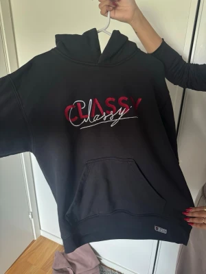 Svart hoodie från Classy med tryck - Svart hoodie från Classy med stor ficka framtill och huva. På bröstet står det 'Classy' i röd och vit brodyr, och på ryggen finns samma text i rött. Hoodien har en avslappnad passform och är tillverkad i mjukt bomullsmaterial. Använd fåtal gånger köpt på limit edition för 1299kr