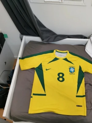 Brasilien fotbollströja Kaká #8 Nike - Gul Brasilien fotbollströja från Nike med gröna detaljer, nummer 8 och Kaká på ryggen. Tröjan har korta ärmar, rund hals och är tillverkad i lätt, ventilerande material. Perfekt för dig som älskar fotboll och vill bära landslagets färger.