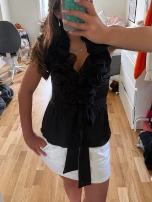 Svart blus med volang och knyt - Supersnygg svart blus med djup v-ringning, stora volangdetaljer framtill och knyt i midjan. Ärmlös modell i ett lätt och luftigt material som ger en elegant look. Perfekt att styla med kjol eller jeans för en trendig outfit. Den saknar en knapp men inget som stör när den sitter på!