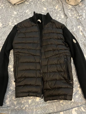 Svart dunväst från Moncler - Snygg svart dunväst från Moncler med stickade svarta ärmar och hög krage. Västen har två dragkedjeförsedda fickor framtill och ett Moncler-märke på ena ärmen. Perfekt för lager-på-lager och streetstyle.