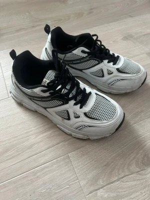 Vita och svarta mesh sneakers - Snygga sneakers i vitt och svart med meshpaneler och svarta detaljer. Skorna har svarta skosnören och en chunky sula för en sportig look. Perfekta för dig som gillar streetwear och vill ha bekväma skor med cool design.