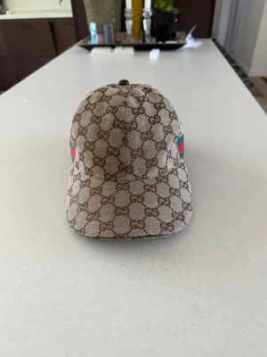 Gucci beige keps med GG-mönster - Säljer en beige keps från Gucci med det klassiska GG-monogrammet över hela kepsen. På sidan finns en grön och röd rand som ger en ikonisk touch. Kepsen har böjd skärm och är tillverkad i canvasmaterial.