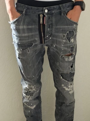 Grå slitna jeans Dsquared2 - Säljer ett par grå jeans från Dsquared2 med slitna detaljer, hål och färgstänk. Jeansen har en unik dragkedja framtill och en avslappnad passform. Märkeslapp bak i midjan och cool streetwear-vibe.
