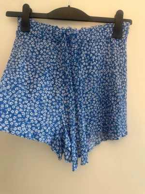 Blå blommiga shorts med snörning strl 38 - Säljer ett par blå shorts med vitt blommönster och snörning i midjan. De har en luftig passform, perfekta för varma dagar. Materialet känns lätt och svalt, vilket gör dem sköna att bära under sommaren.