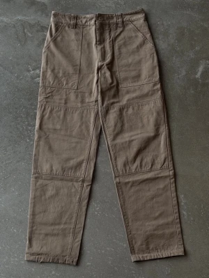 Axel Arigato Double Knee Carpenter Pants – Brun (M) - Bruna arbetsbyxor i kraftig bomullscanvas från Axel Arigato. Modellen har förstärkta paneler över knäna (double knee), hammarhank samt verktygsficka på höger ben. Rak, rymlig passform. Nypris ca 2300 kr.  OBS: Se sista bilden då ena benöppningen har fransat sig.