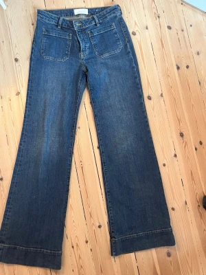 Blåa Jeanerica Jeans 28/34 - Säljer ett par blå jeans med hög midja och utsvängda ben. Jeansen har stora framfickor och klassisk femficksdesign. Materialet är denim i bomull och färgen är mörkblå. Perfekta för en trendig och avslappnad stil. 