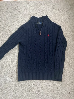 Mörkblå kabelstickad tröja Polo Ralph Lauren - Mörkblå kabelstickad tröja från Polo Ralph Lauren med halv dragkedja och röd broderad logga på bröstet. Tröjan har ribbade muddar och hög krage, perfekt för lager på lager. Klassisk och tidlös design i mjukt material.