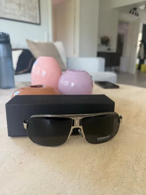 Svarta solglasögon från Porsche Design - Snygga solglasögon från Porsche Design med svarttonade glas och silverfärgad metallbåge. Klassisk och sportig design med markerad brygga och logga på linsen. Perfekta för dig som vill ha en stilren accessoar med exklusiv känsla.