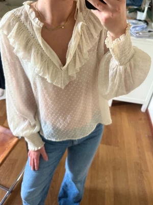 Vit blus med volang - Säljer en vit, transparent blus med volanger längs v-ringningen och axlarna. Blusen har ett diskret prickigt mönster och breda, långa ärmar med smockade muddar. Strl XL men liten i storlek så skulle säga att det är som en M