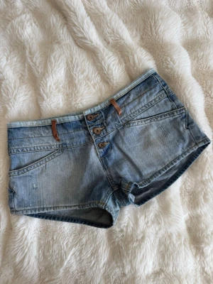 Blå denimshorts med knappar - Snygga blå denimshorts med låg midja och tre knappar framtill. Shortsen har slitna detaljer, bruna bälteshällor och klassiska fickor både fram och bak. Perfekta för varma dagar och en avslappnad stil. Passar S-M