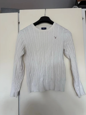 Vit kabelstickad tröja från GANT - Säljer en vit kabelstickad tröja från GANT med rund hals och broderad logotyp på bröstet. Tröjan har långa ärmar och ribbade muddar vid ärmslut och nederkant. Perfekt för dig som gillar klassisk och stilren design.