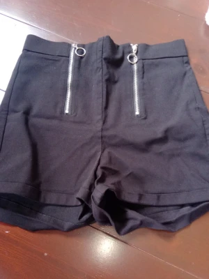 Svarta högmidjade shorts med dragkedjor - Snygga svarta högmidjade shorts med två silverfärgade dragkedjor framtill. Shortsen har en clean look och är tillverkade i ett mjukt bomullsmaterial. Perfekta för dig som gillar en modern och edgy stil.