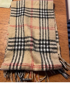 Beige rutig halsduk från Burberrys - Klassisk beige halsduk från Burberrys i 100% lammull med det ikoniska rutmönstret i svart, vitt och rött. Halsduken har fransar i ändarna och är tillverkad i England. Perfekt accessoar för att lyfta din outfit.
