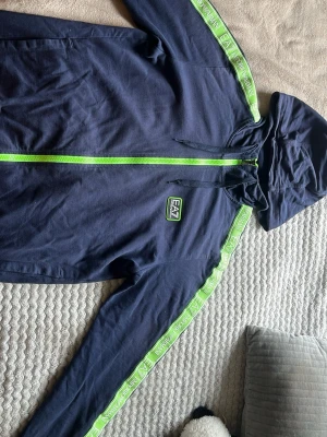 Marinblå EA7 hoodie med gröna detaljer - Snygg marinblå hoodie från EA7 Emporio Armani med huva och dragkedja i neon-grönt. Grön logga på bröstet och gröna band med EA7- och Armani-tryck längs ärmarna. Tillverkad i mjukt bomullsmaterial och har fickor på sidorna. Perfekt för en sportig och trendig look.
