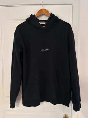 Svart hoodie från Saint Laurent - Snygg svart hoodie från Saint Laurent med minimalistisk vit logga på bröstet. Klassisk passform med huva och stor magficka framtill. Tillverkad i mjukt material som känns skönt mot huden. Perfekt för en avslappnad och trendig look. 