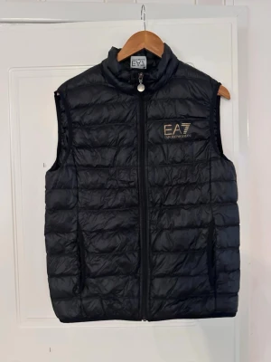 Svart dunväst från EA7 Emporio Armani - Snygg svart dunväst från EA7 Emporio Armani med quiltad design och dragkedja framtill. Västen har hög krage och logga på bröstet. Perfekt för lager på lager och ger en sportig vibe. Materialet är lätt och smidigt, vilket gör den enkel att bära. Sitter som S