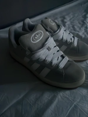 Adidas gråvita sneakers med gummisula - Snygga Adidas sneakers i grått och vitt med klassiska tre ränder på sidan och stor logga på plösen. Skorna har breda vita skosnören och en slitstark gummisula med fiskbensmönster. Perfekta för dig som gillar retrostil och streetwear.