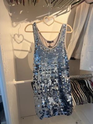 Silver paljettklänning från Tsega - Säljer en glittrig silverfärgad klänning från Tsega Fashion Collection. Klänningen är täckt av stora paljetter, har ärmlös design och rund halsringning. Perfekt för dig som vill sticka ut och glänsa på nästa fest.