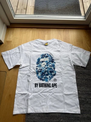 Vit BAPE t-shirt med blått camomönster - Säljer en clean vit t-shirt från BAPE med det ikoniska apansiktet i blått camomönster på bröstet och texten 'BY BATHING APE' under. Klassisk passform och korta ärmar, perfekt till streetwear-stilen.