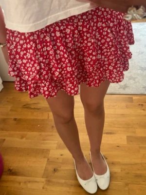 Röd blommig volangkjol - Supersöt röd kortkjol med vitt blommönster och volangkant. Kjolen har en luftig och flowy passform som ger en skön sommarkänsla. Perfekt att matcha med en enkel topp för en trendig look.