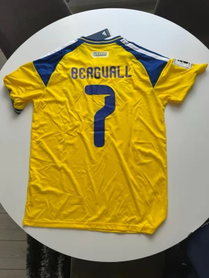 Sverige matchtröja Bergvall #7 Adidas - Gul och blå matchtröja från Adidas för Sveriges landslag med Bergvall och nummer 7 på ryggen. Tröjan har korta ärmar, broderad landslagssköld och Adidas-logga framtill samt CLIMACOOL-material för bra ventilation. Perfekt för fotbollsfans.
