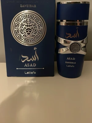 Lattafa Asad Zanzibar parfym - Lattafa Asad Zanzibar parfym i en elegant mörkblå flaska med silverdetaljer och orientaliskt mönster. Flaskan har en unik form med metallband och dekorativ medaljong. Förpackningen matchar flaskans design och ger en lyxig känsla.